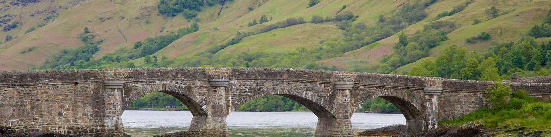 A stone bridge over a body of water with hills in the background.