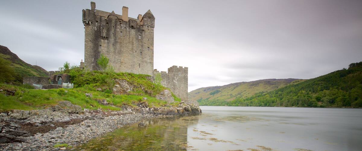 A castle by a calm river with surrounding hills.