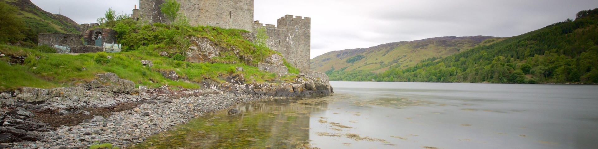 A castle by a calm river with surrounding hills.