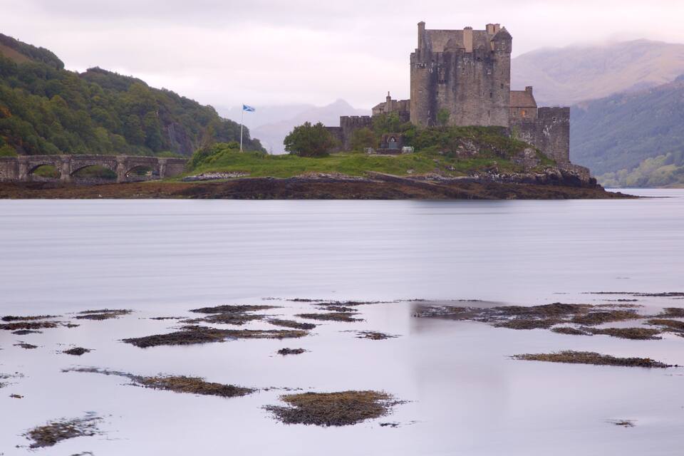 A castle on a hill overlooking a calm lake, surrounded by mountains.