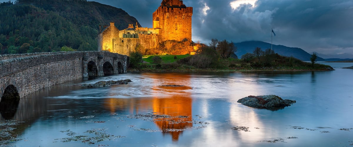 A historic castle by a calm river with a stone bridge, surrounded by hills and under a cloudy sky.