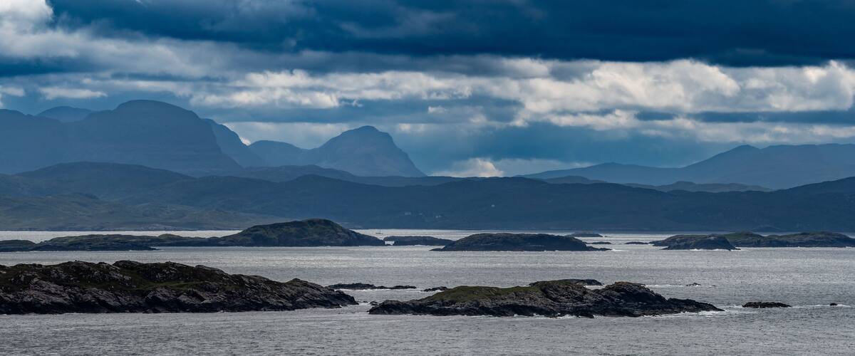 A coastal landscape with rocky outcrops and distant mountains under a cloudy sky.
