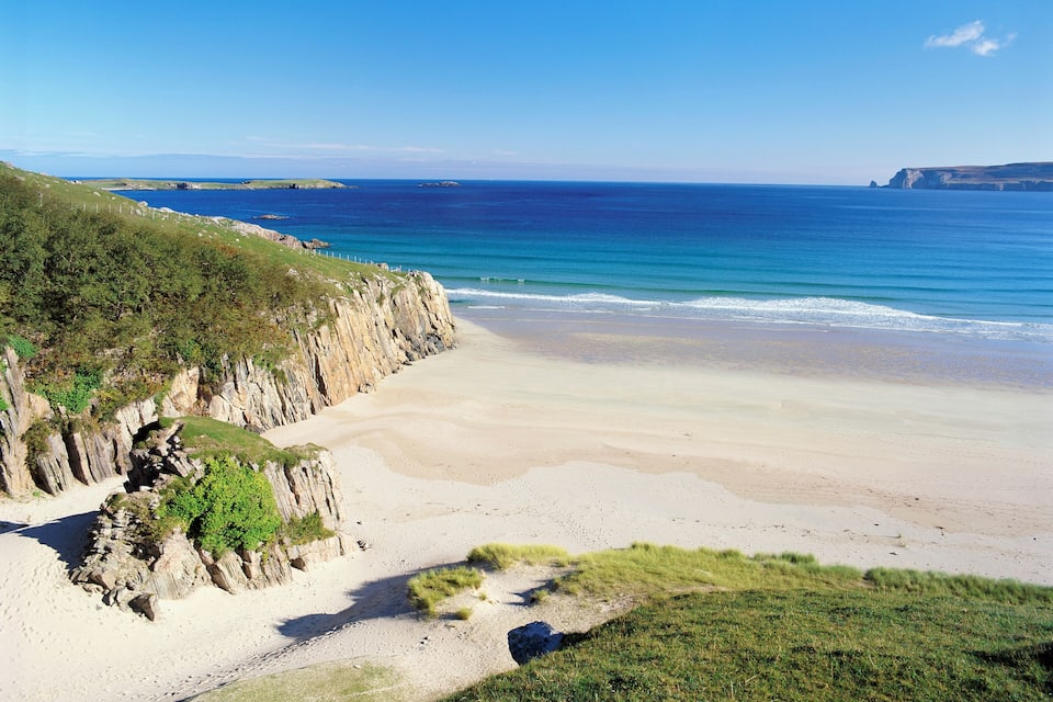 A coastal landscape with a sandy beach, grassy cliffs, and a clear blue sea.