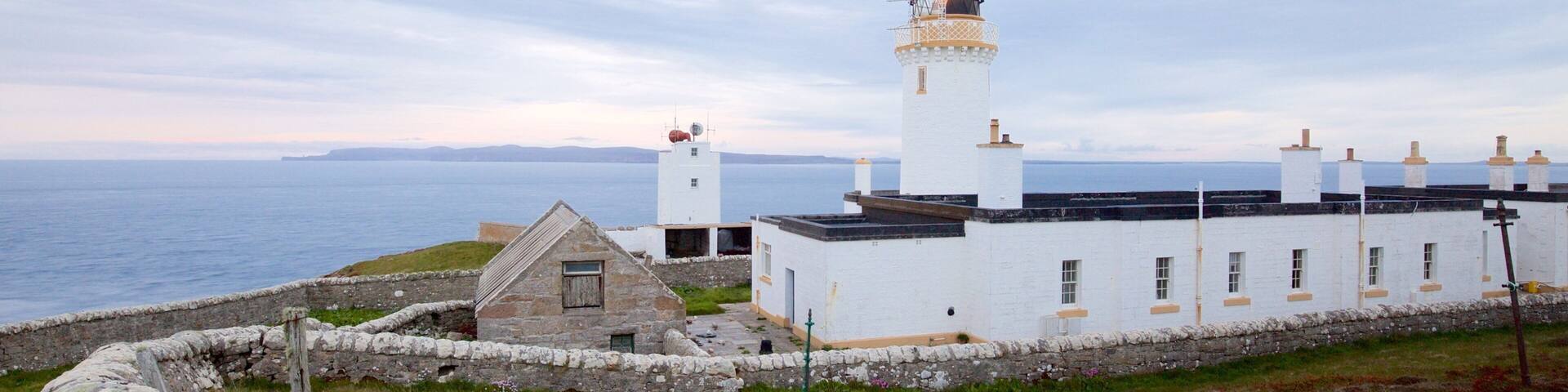 A lighthouse with a white building beside it, a stone wall, and the sea in the background.