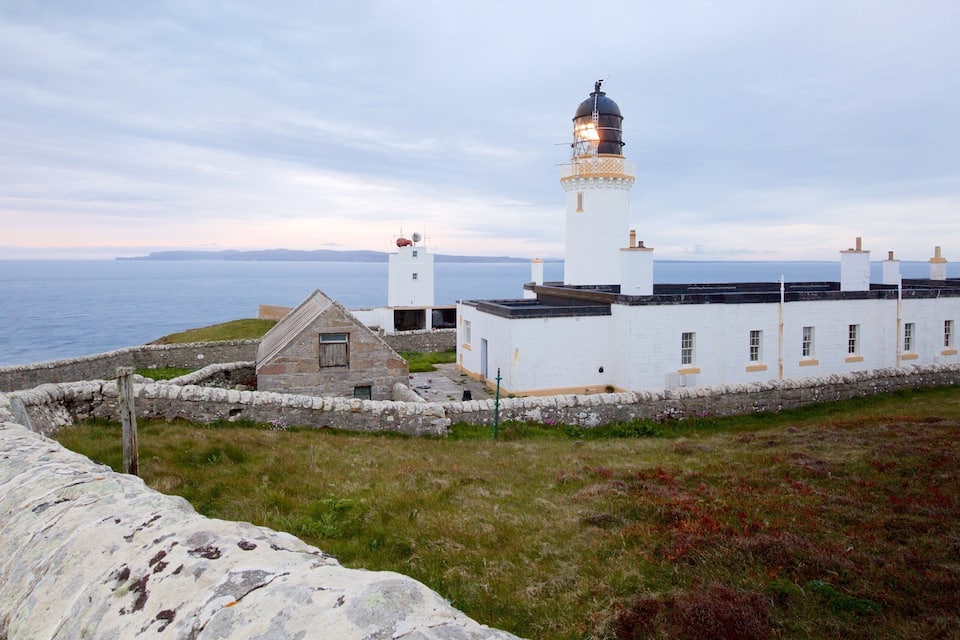 Un faro con un edificio blanco a su lado, un muro de piedra y el mar al fondo.