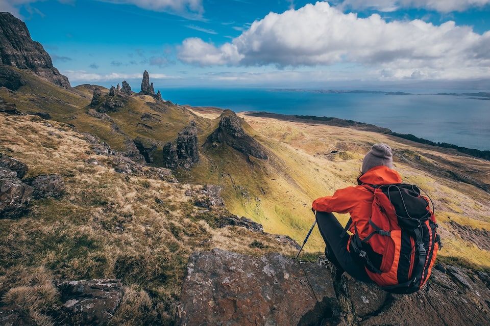 A hiker in a red jacket and gray beanie sitting on a rock, overlooking a mountainous landscape with a body of water in the distance.