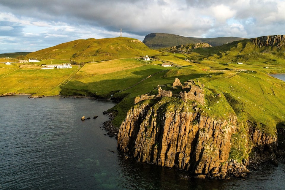 A coastal landscape with a rocky outcrop, grassy hills, and a small white building.