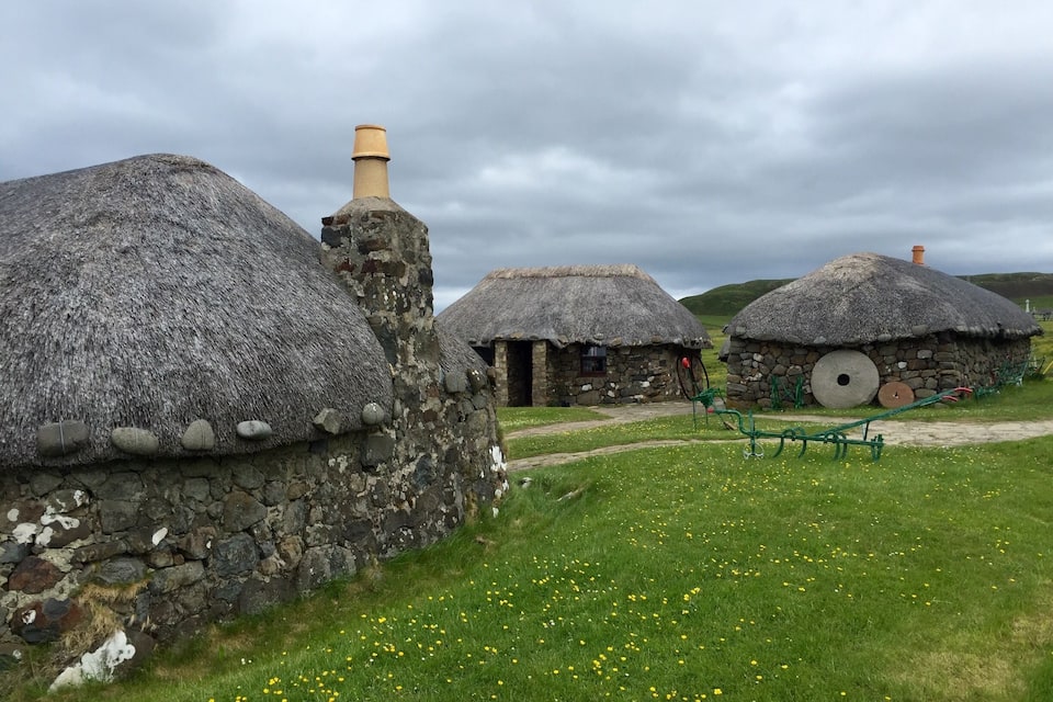 Thatched-roof stone houses with thatched roofs, a stone chimney, and a green tractor in a grassy field.