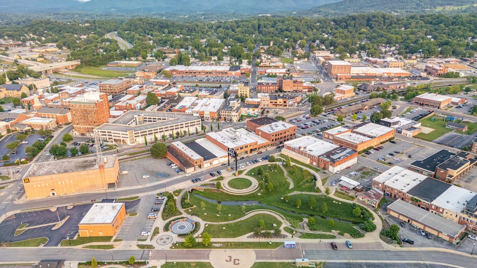 An aerial view of a city with a central green park, a large building with a 'J' on its roof, and surrounding residential and commercial buildings.