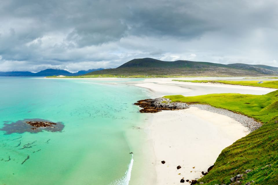 A coastal landscape with a sandy beach, clear turquoise waters, and green hills under a cloudy sky.