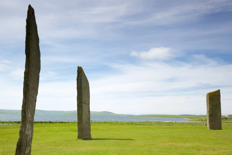 Three standing stones in a grassy field with a lake and hills in the background.