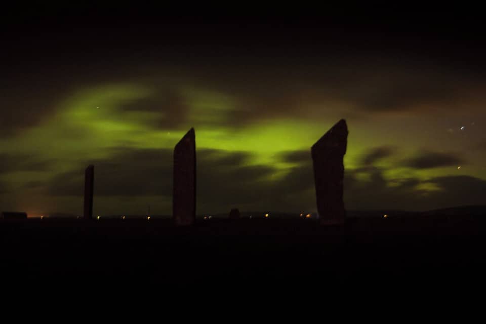 Tres piedras verticales contra un cielo nocturno con aurora verde.