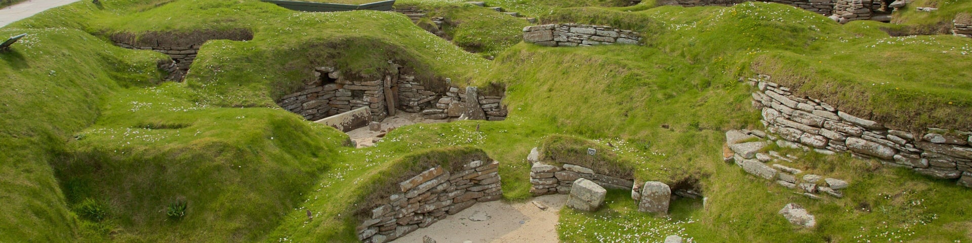 A grassy archaeological site with stone structures and a flagpole.
