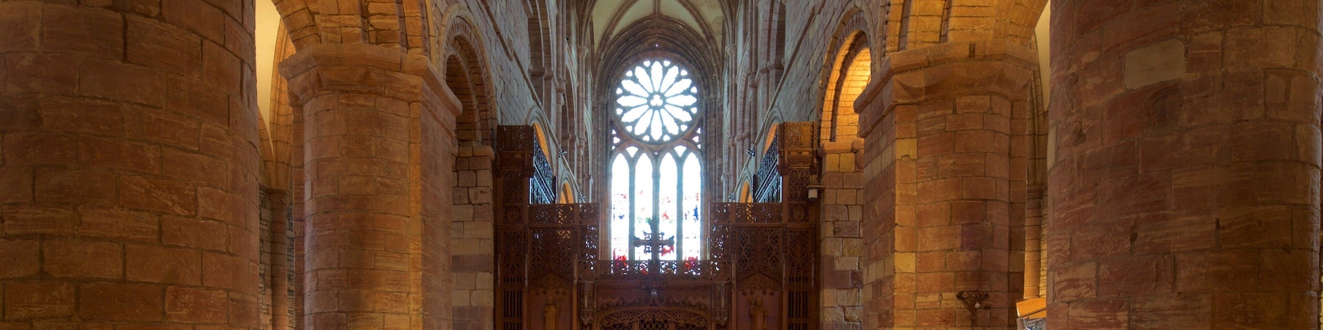 Interior de uma catedral com altos tetos abobadados, grandes janelas de vitrais e bancos de madeira ornamentados.