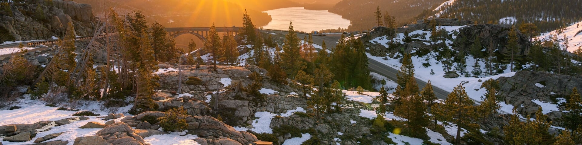 A mountainous landscape with a river, snow-covered peaks, and a bridge.