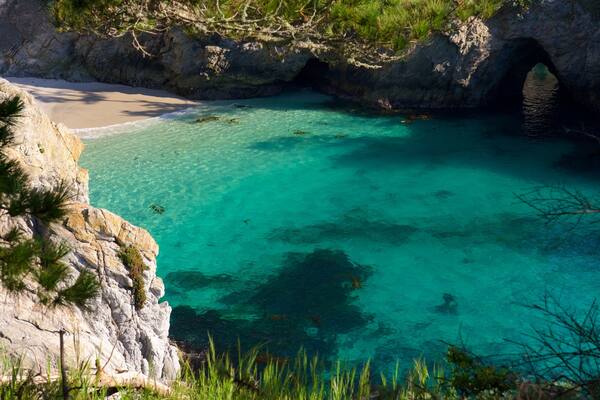 Ein türkisfarbenes Meer mit klarem Wasser, ein Sandstrand und felsige Klippen mit Vegetation.