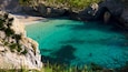 A turquoise sea with clear water, a sandy beach, and rocky cliffs with vegetation.