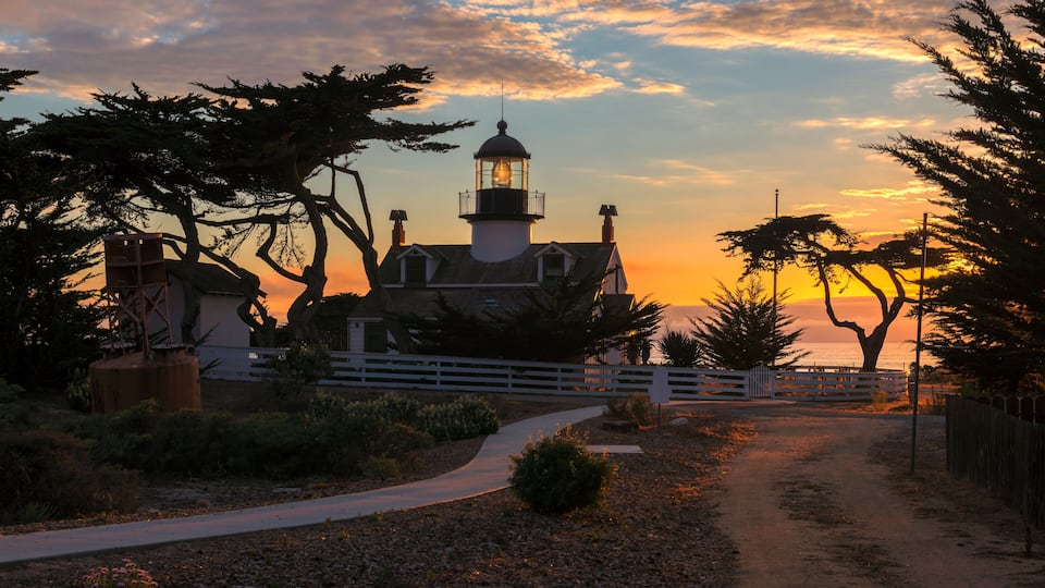 A lighthouse at sunset with a white fence and trees in the foreground.