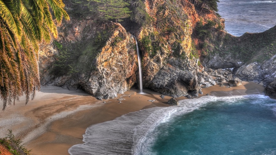 A coastal landscape with a waterfall, sandy beach, and rocky cliffs.