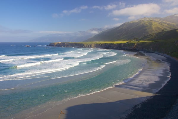 A coastal landscape with a sandy beach, ocean waves, and a mountainous backdrop.
