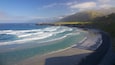 A coastal landscape with a sandy beach, ocean waves, and a mountainous backdrop.