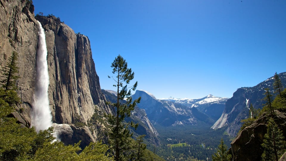 A waterfall cascading down a steep cliff, surrounded by dense forest and snow-capped mountains under a clear blue sky.