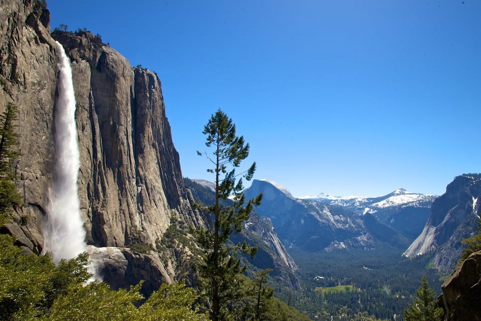 A waterfall cascading down a steep cliff, surrounded by dense forest and snow-capped mountains under a clear blue sky.