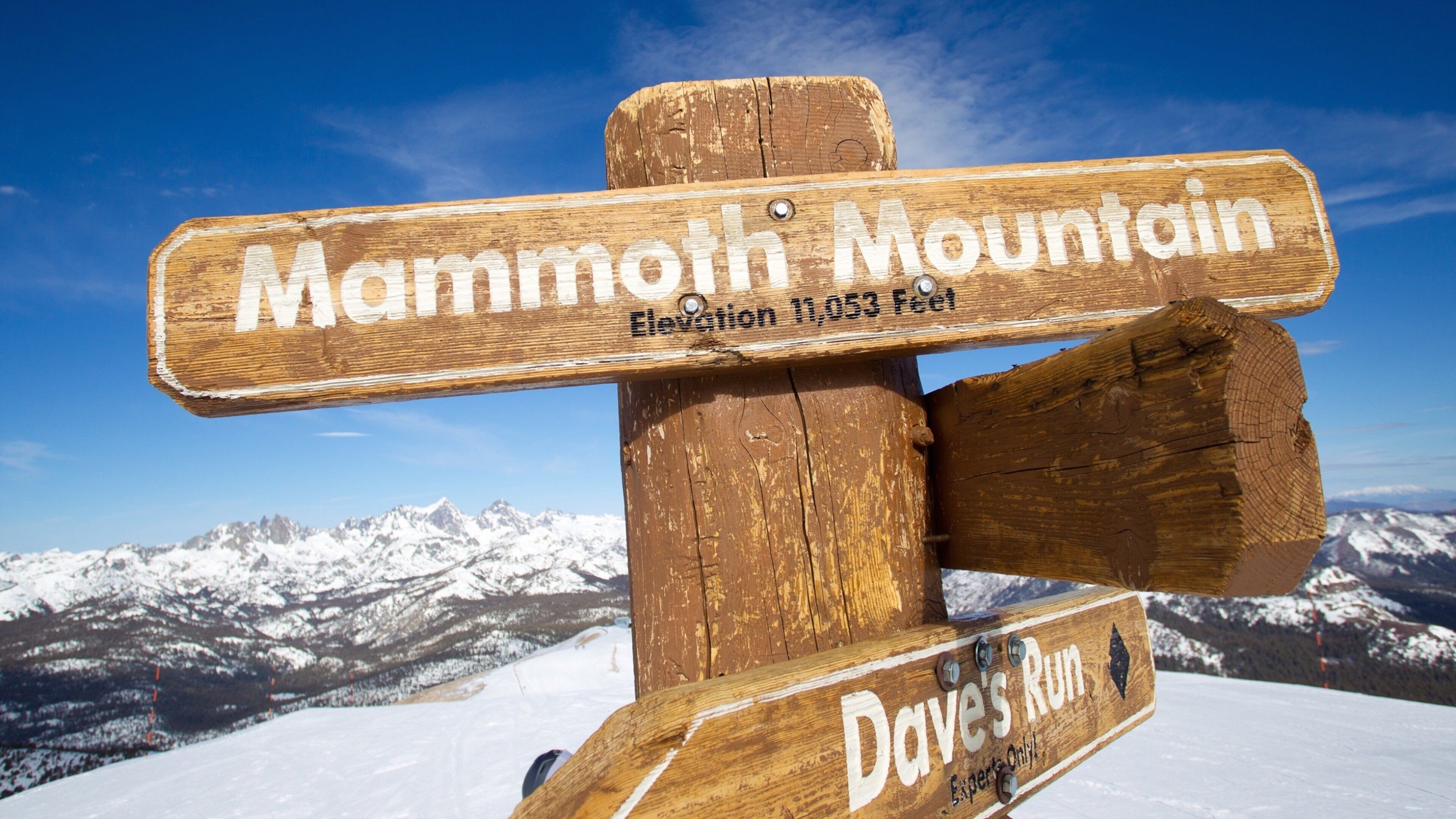 Ein hölzernes Wegweiser mit der Aufschrift 'Mammoth Mountain' und 'Höhe 3.370 Meter', vor einer verschneiten Berglandschaft.