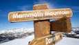 Ein hölzernes Wegweiser mit der Aufschrift 'Mammoth Mountain' und 'Höhe 3.370 Meter', vor einer verschneiten Berglandschaft.