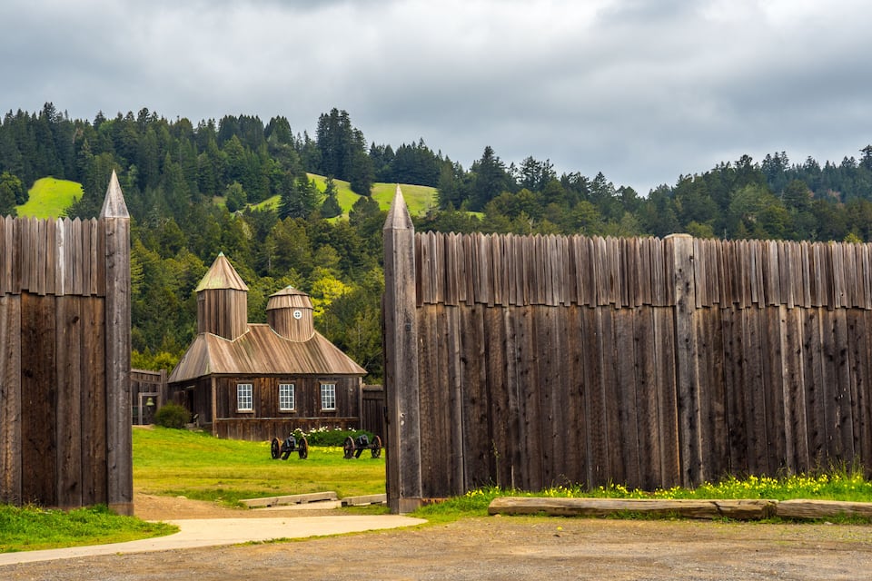 A rustic wooden fence, a small building with a bell tower, and a green hill in the background.
