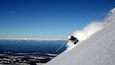 Un skieur dévalant une pente abrupte recouverte de neige, avec un ciel bleu dégagé et des nuages lointains en arrière-plan.