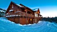 A wooden lodge with a balcony and snow-covered roof, surrounded by a snowy landscape.