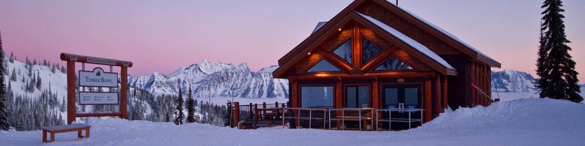 A wooden lodge with a glass facade, surrounded by snow-covered mountains and a wooden signboard.