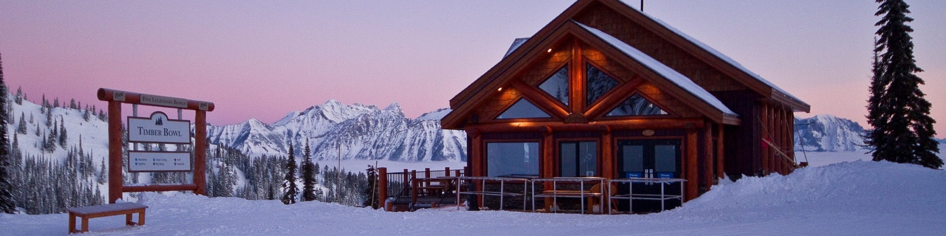 A wooden lodge with a glass facade, surrounded by snow-covered mountains and a wooden signboard.