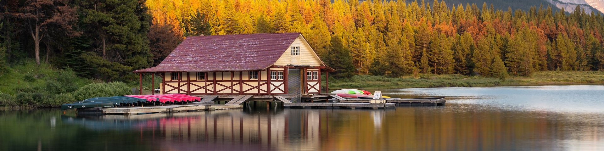 Una cabaña junto a un lago tranquilo, con follaje otoñal y montañas al fondo.