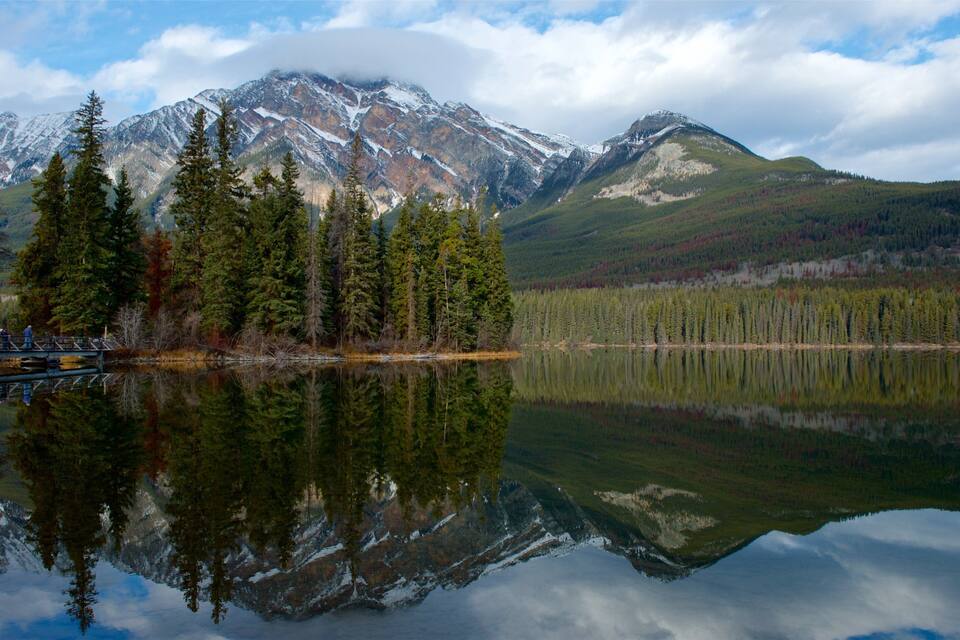A mountain range with snow-capped peaks, a calm lake reflecting the trees, and a wooden bridge in the foreground.