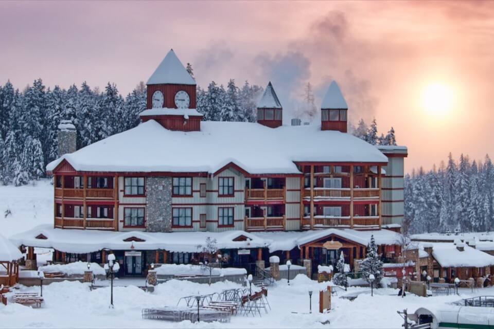 A large wooden lodge with multiple chimneys and a snow-covered roof, surrounded by a snowy landscape and dense trees.