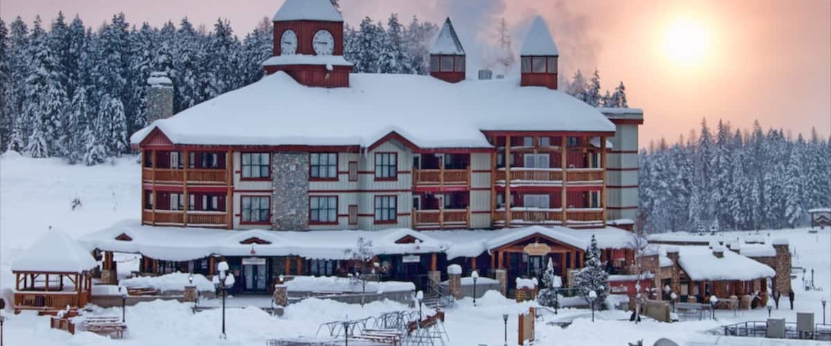 A large wooden lodge with multiple chimneys and a snow-covered roof, surrounded by a snowy landscape and dense trees.