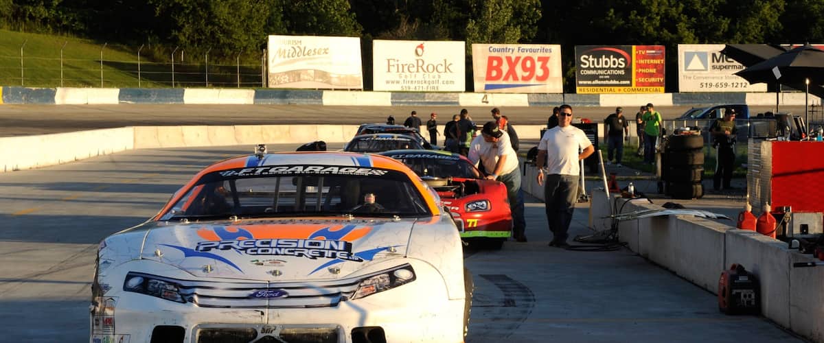A race track with a white and orange race car in the foreground and several people in the background.