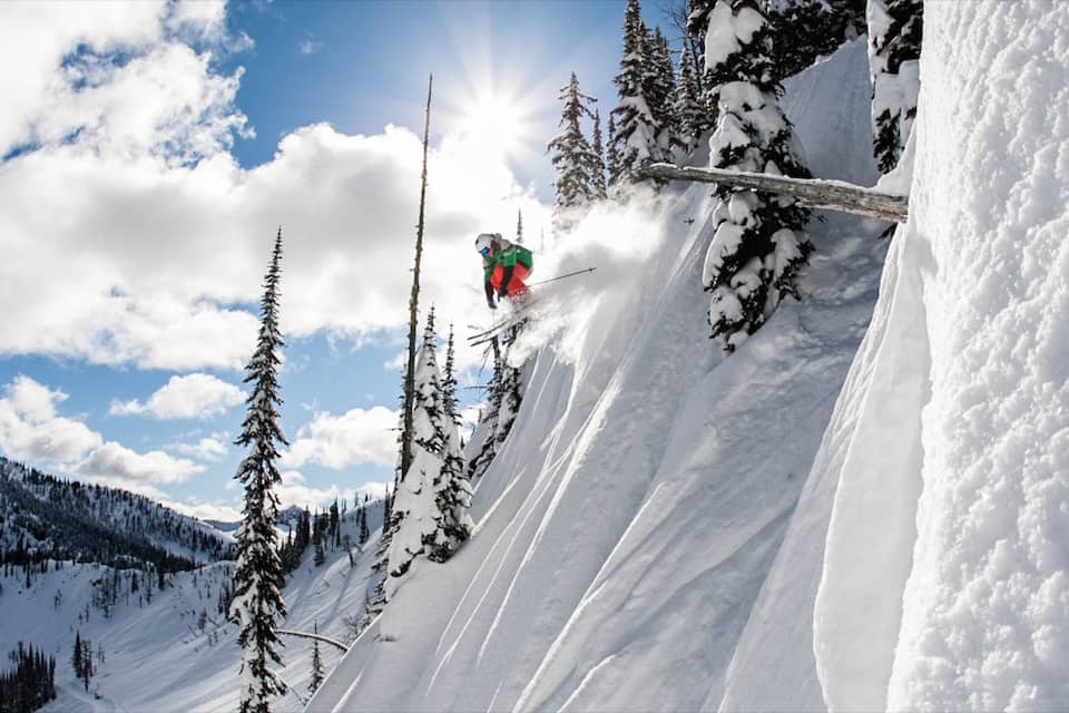 A skier performing a jump on a snowy mountain slope with trees and a clear sky.
