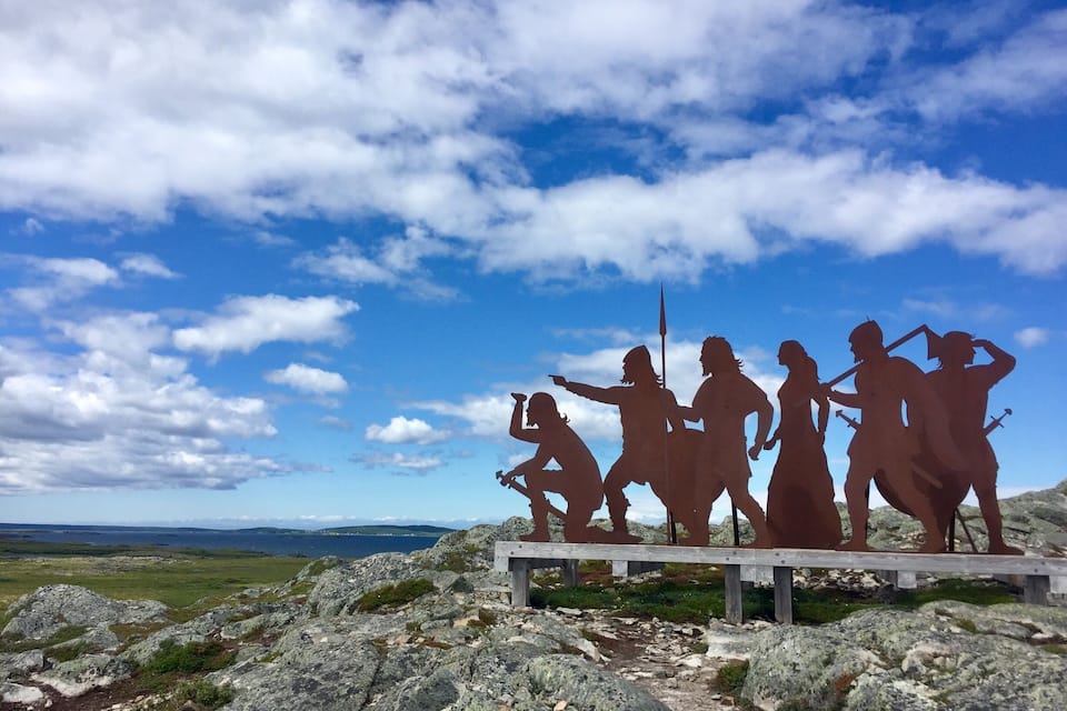 A sculpture of warriors on a wooden platform with a mountainous landscape and a blue sky in the background.