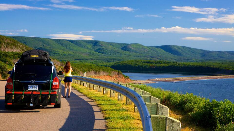 A woman walking along a coastal road with a car parked on the side.