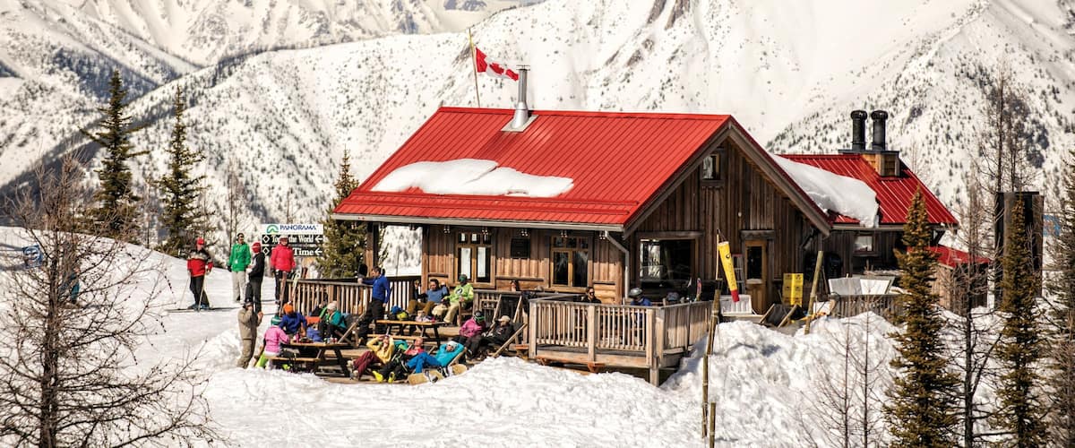 A wooden cabin with a red roof and a Canadian flag, surrounded by snow-covered mountains and trees.