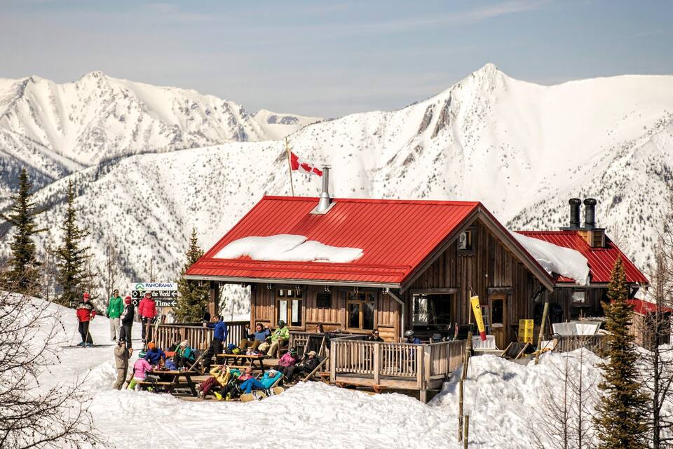 Una cabaña de madera con techo rojo y una bandera canadiense, rodeada de montañas y árboles cubiertos de nieve.