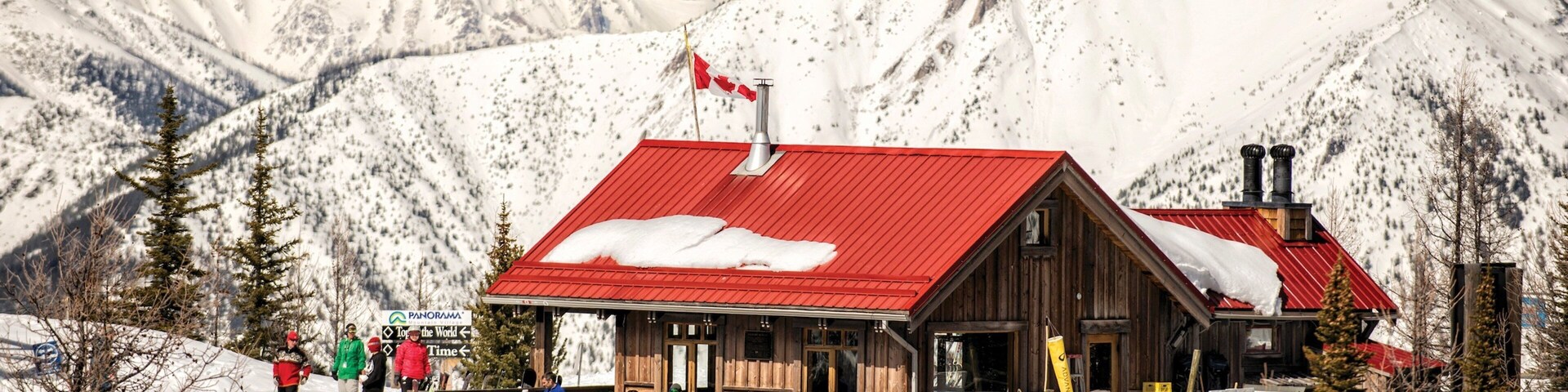 Uma cabana de madeira com telhado vermelho e uma bandeira do Canadá, rodeada por montanhas e árvores cobertas de neve.
