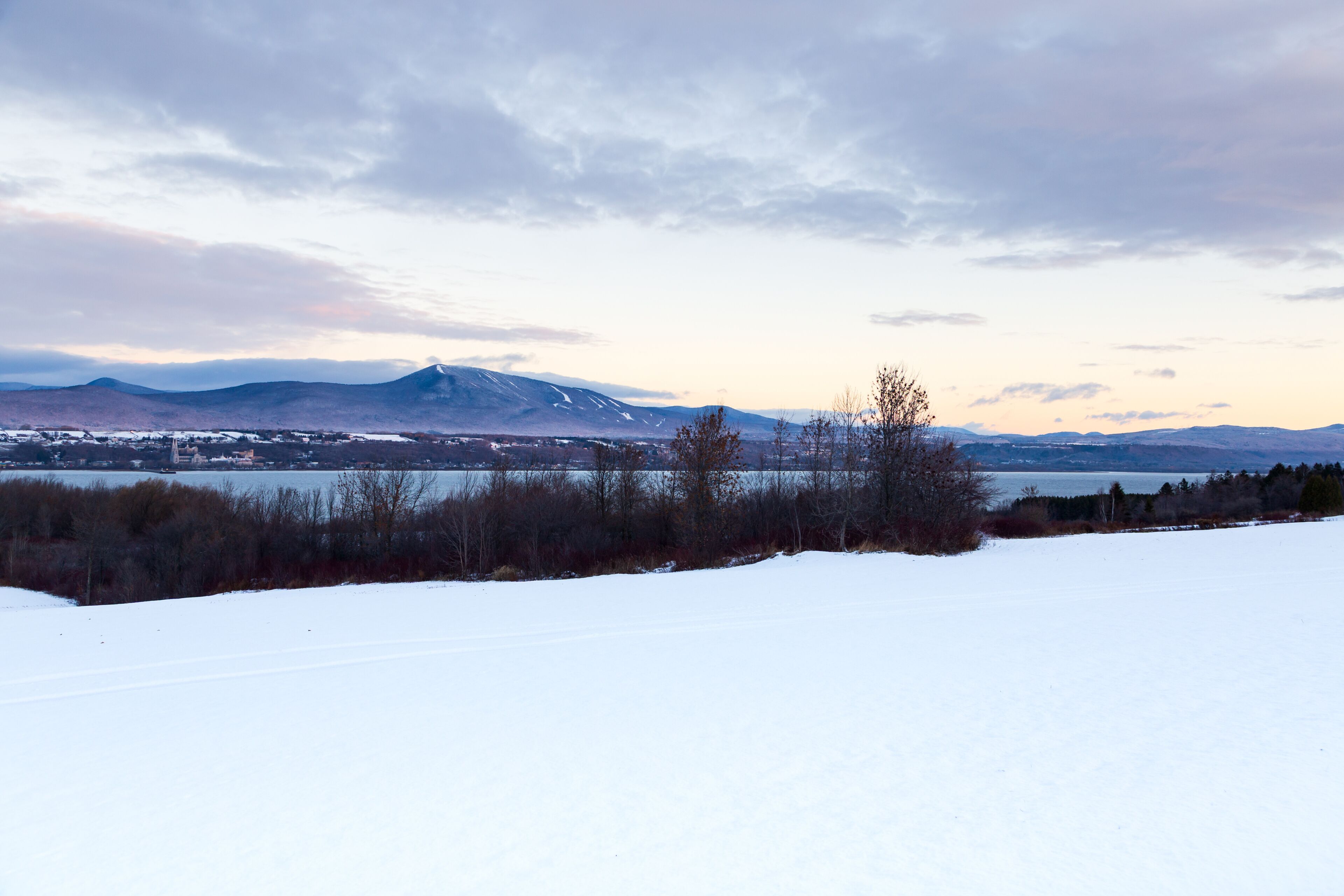 Rural winter landscape seen during a partly cloudy dawn in Saint-François-de-l'Île-d'Orléans, with the St. Lawrence river and Mont Sainte-Anne in soft focus background, Quebec, Canada