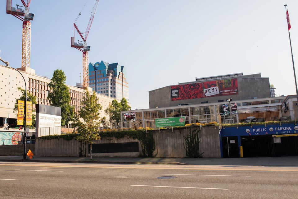 Un carrer de ciutat amb un teatre, un pàrquing públic i algunes grues de construcció.