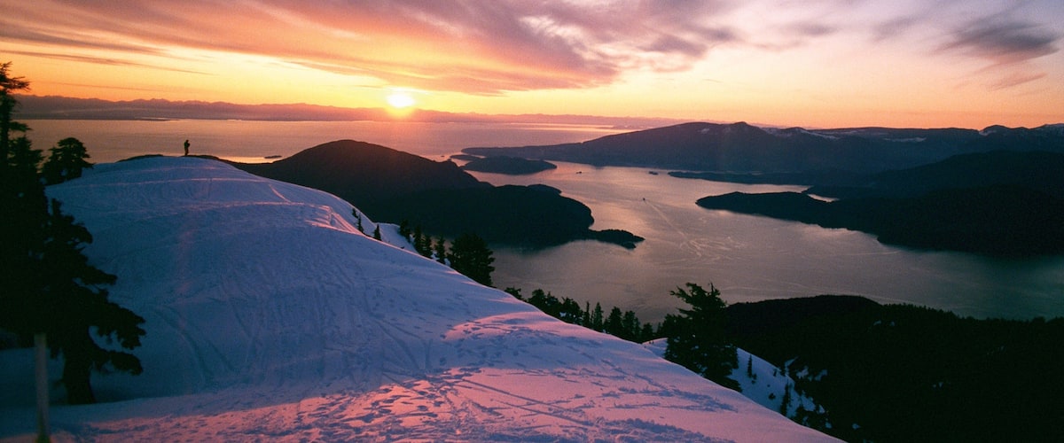 A snow-covered mountain slope with a sunset over a lake and surrounding hills.