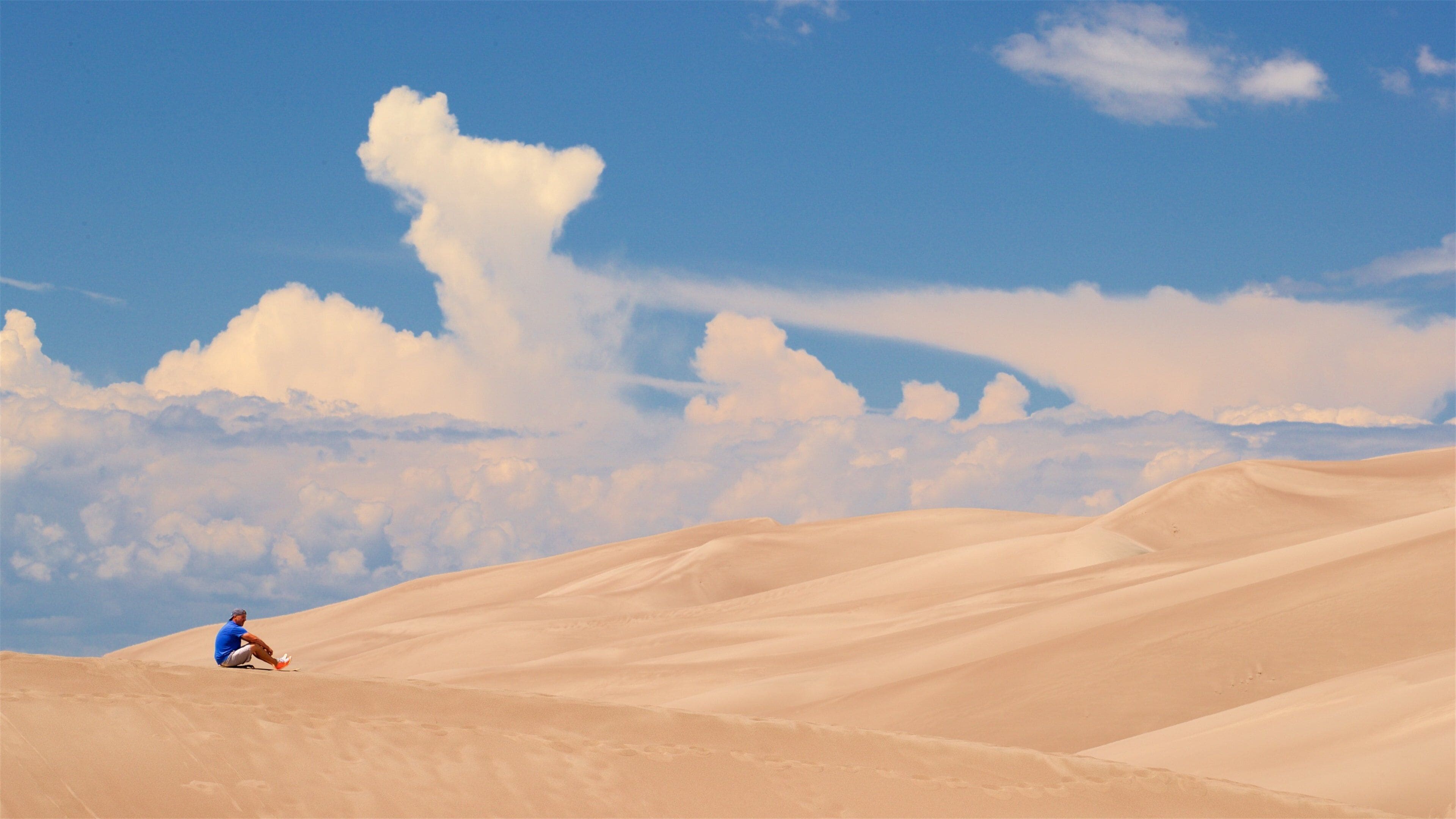 A person sitting on a sand dune with a clear blue sky and clouds in the background.