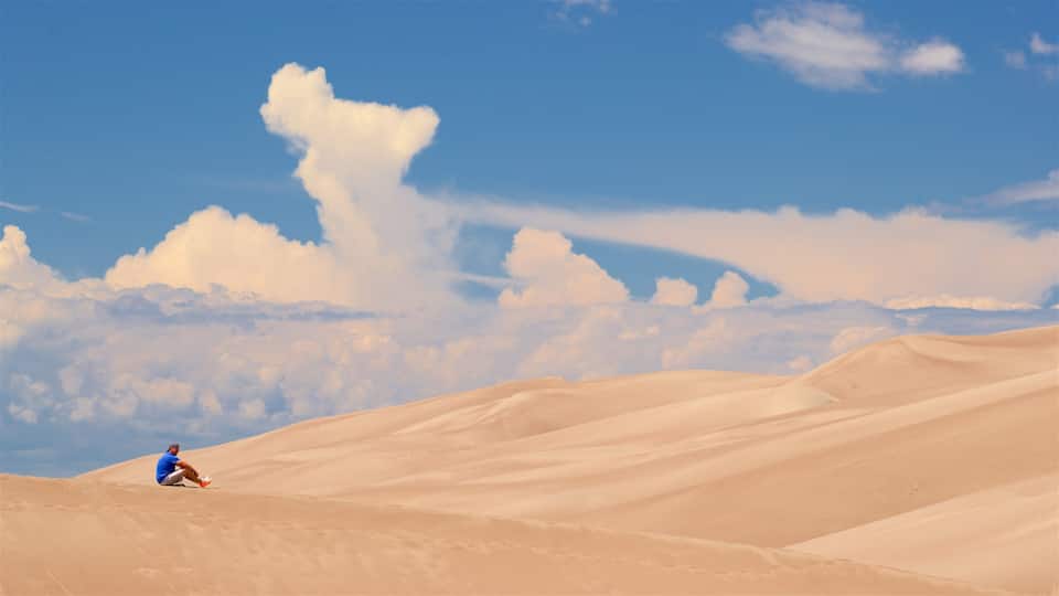 A person sitting on a sand dune with a clear blue sky and clouds in the background.
